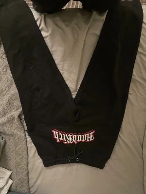 Svarta mjukisbyxor Hoodrich - Svarta mjukisbyxor från Hoodrich med tryck i vitt och rött på ena benet och textdetaljer längs det andra benet. Byxorna har snörning i midjan och muddar vid bensluten. Perfekta för en chill och avslappnad stil.