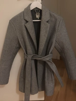 Grå kappa med bälte från ONLY - Stilren grå kappa från ONLY med klassisk krage och knytskärp i midjan. Här är Perfekt för dig som gillar en enkel och tidlös look.
