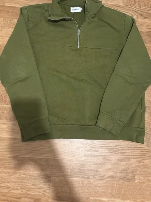 Olivgrön halfzip från East West - Säljer en olivgrön sweatshirt med halv dragkedja från East West. Tröjan har hög krage, långa ärmar och ribbade muddar. Perfekt för en avslappnad och stilren look. Materialet är mjukt och bekvämt, troligen bomull eller bomullsblandning.