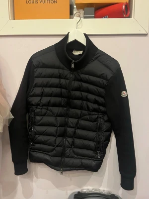 Svart quiltad tröja från Moncler - Snygg svart tröja från Moncler med quiltad front och ärmar i mjukt tyg. Tröjan har dragkedja framtill, två fickor med dragkedja och Moncler-logga på ärmen. Perfekt för dig som gillar exklusiv streetwear och vill ha något stilrent.