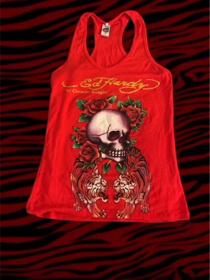 Röd Ed Hardy linnetopp med tryck - Säljer en röd linnetopp från Ed Hardy med stort tryck av dödskalle, rosor och tigrar på framsidan. Toppen har brottarrygg och logga i guldtext både fram och bak. Perfekt för dig som gillar statement-plagg och edgy stil. (Några blind har ramlat av) 