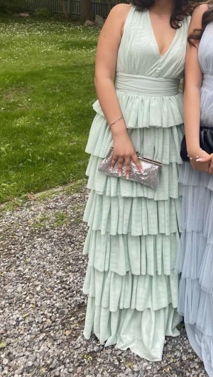  balklänning med volanger - Superfin långklänning i mintgrönt med djup v-ringning och markerad midja. Klänningen har flera lager volanger som ger en riktigt drömmig och flowy känsla. Perfekt för dig som vill sticka ut med en romantisk och elegant look.