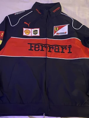Svart Ferrari jacka  - Cool svart jacka från Ferrari med röda och vita detaljer, logotyper och broderade märken från bland annat Shell och UPS. Jackan har dragkedja framtill och sportig design, perfekt för dig som gillar motorsport och snygga streetwear-plagg. Strl.M men bassar både L samt XL. Kan gå ner i pris vid snabb affär