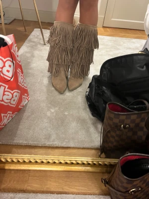 Isabel marant liknande  - Säljer ett par beiga cowboy boots med massor av fransar och spetsig tå. Bootsens material är mocka och de har en klassisk westernstil som sticker ut. Perfekta för dig som vill ha något unikt och trendigt till din outfit. HELT OANVÄNDA 