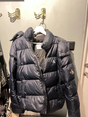 Mörkblå pufferjacka från Calvin Klein - Mörkblå pufferjacka från Calvin Klein med huva och dragkedja. Jackan har CK-logga på bröstet och ärmen, samt två sidofickor. Perfekt för kalla dagar och har en klassisk, stilren look. Varsamt använd 