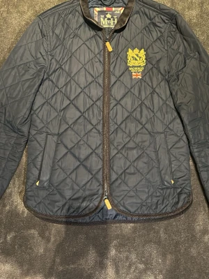 Marinblå quiltad vindjacka Morris - Stilren marinblå vindjacka från Morris med quiltat mönster och guldfärgat broderi på bröstet. Jackan har dragkedja, två fickor framtill och detaljer i brunt vid dragkedja och nederkant. Perfekt för en klassisk och sportig look.