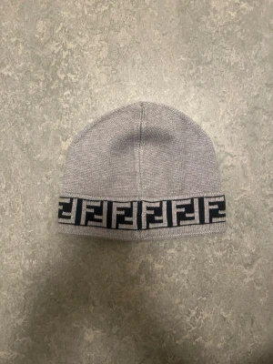 Fendi Mössa grå - Snygg grå stickad mössa från Fendi med svart och grått logomönster runt kanten. Perfekt accessoar för att lyfta din outfit och hålla dig varm under kalla dagar. Ny med tag