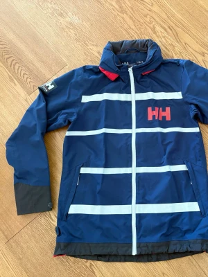 Blå vindjacka från Helly Hansen stl 176/XS - Blå vindjacka från Helly Hansen med vita reflexränder och röd HH-logga på bröstet. Jackan har hög krage, röd huva och dragkedja framtill. Tillverkad i slitstark polyester och har lång ärm med svarta detaljer vid ärmslut och nederkant.