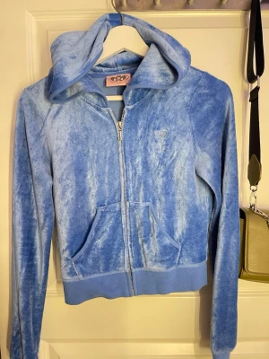 Blå velourhoodie från Juicy Couture - Snygg blå velourhoodie från Juicy Couture med dragkedja framtill, huva och två fickor. Ikoniskt JC-broderi på bröstet och ribbade muddar vid ärmslut och nederkant. Perfekt för en avslappnad och trendig look.