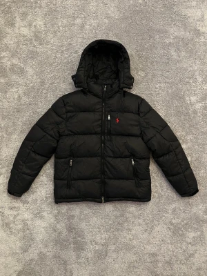 Polo Ralph Lauren dunjacka  - Svart dunjacka från Polo Ralph Lauren med huva och dragkedja framtill. Jackan har två fickor med dragkedja och klassisk röd logga på bröstet. Perfekt för kalla dagar med sin quiltade design och varma fyllning.