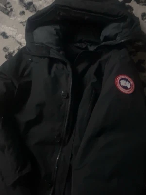 Svart dunjacka från Canada Goose - Säljer en svart dunjacka från Canada Goose med klassisk patch på ärmen och huva. Jackan har dragkedja och knappar framtill, perfekt för kalla vinterdagar. Stilsäker och tidlös design som håller dig varm hela säsongen. Hjälper en vän storlek L men passar M perfekt 