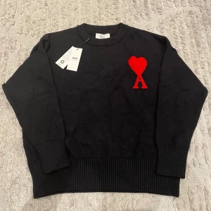 Svart stickad tröja från Ami Paris - Säljer en svart stickad tröja från Ami Paris med deras ikoniska röda hjärta och bokstaven A broderat på bröstet. Tröjan har rund hals, ribbade muddar och är tillverkad i mjukt material. Perfekt för dig som gillar stilrena plagg med en twist.