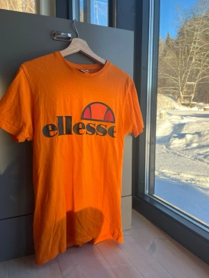 Orange t-shirt från Ellesse strl S - Snygg orange t-shirt från Ellesse med stort tryckt logotyp på bröstet. Klassisk rund halsringning och korta ärmar. Perfekt för dig som gillar sportig och färgstark stil.