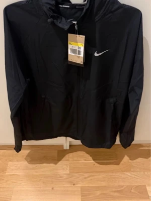Nike men jogging jacket - Snygg svart vindjacka från Nike med huva och dragkedja framtill. Jackan har en vit Nike-logga på bröstet och praktiska fickor. Perfekt för blåsiga dagar och sportiga outfits. Köpt direkt på Nikes hemsida för 999kr, säljer för 450kr 💕