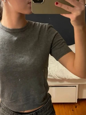 Grå croppad t-shirt från H&M - En enkel och stilren grå t-shirt från H&M. Modellen är croppad med ribbade kanter vid ärmslut och nederkant. T-shirten har rund halsringning och är tillverkad i mjuk bomull, perfekt för en avslappnad look.