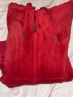 Röd velour hoodie från Juicy Couture - Säljer en röd zip-up hoodie från Juicy Couture i mjuk velour. Hoodien har huva med dragsko, ribbade muddar och fickor framtill. Juicy Couture-logga broderad på bröstet och snygga metalldetaljer på snörena. Perfekt för en chill och trendig look. 