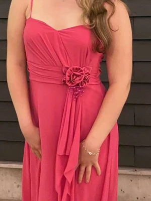 Rosa långklänning med blomdetalj - Elegant rosa långklänning med smala axelband och v-ringad urringning. Klänningen har en dekorativ blomma i midjan med pärldetaljer och draperad tygdetalj framtill. Perfekt för bal eller festliga tillfällen. Endast använd 1 gång på en bal. Skulle säga att den passar många storlekar från XS-M. 