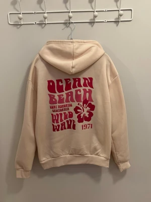 Hoodie - Snygg beige/ljusgul hoodie med stor mörkrosa retroinspirerad text och hibiskusblomma på ryggen samt mindre blomma på bröstet. Har huva med dragsko och en stor magficka. Texten på ryggen lyder 'Ocean Beach Wild Wave 1971'. Perfekt för en chill och avslappnad stil.