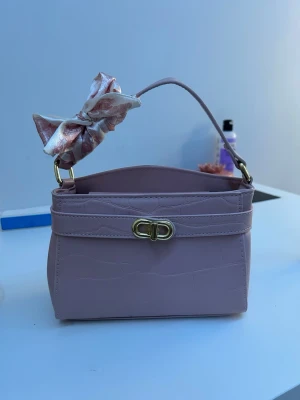 Rosa handväska med rosett och axelrem - Snygg ljusrosa handväska i syntet med guldfärgat spänne och dekorativ rosett på handtaget. Väskan har en avtagbar axelrem och stilren, boxig form. Perfekt accessoar för att lyfta din outfit.