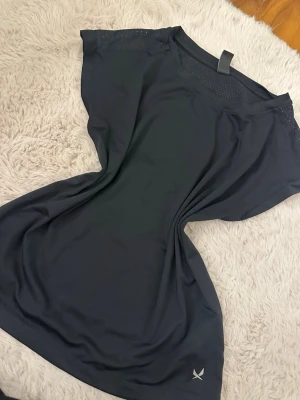 Svart sportig topp med meshdetaljer - Svart sportig topp f med meshdetaljer vid axlarna och rund halsringning. Tillverkad i ett lätt och stretchigt syntetmaterial som passar perfekt för träning. Liten logga nere vid fållen och korta ärmar för en avslappnad look.