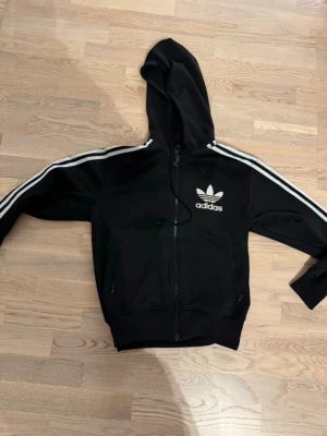 Adidas tröja med dragkedja vintage  - Adidas vintage tröja med dragkedja.  Svart tröja med vita streck.  Storlek : S , kort passform.  Inga defekter , lite skav på hänget till dragkedjan se bild nummer 6.