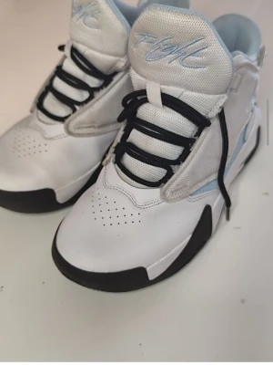 Nike Jordan sneakers vit/blå - Snygga Nike Jordan sneakers i vitt och ljusblått med svarta detaljer. Skorna har svart snörning, rund tå och perforerade partier för ventilation. Tillverkade i syntetmaterial och skinn med klassisk 'Flight'-logga på plösen och Jumpman-logga på sidan.