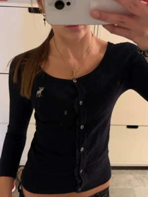 Mörkblå kofta Abercrombie & Fitch - Säljer en mörkblå, figurnära 3/4 ärmad kofta från Abercrombie & Fitch med silverfärgade knappar och deras klassiska älg-logga broderad på bröstet. Barnstorlek XL passar en vanlig XS. Koftan har trekvarts ärmar och ribbade muddar. Perfekt att styla med jeans eller kjol.