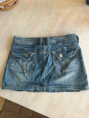 Blå lågmidjad jeanskjol  - Blå lågmidjad jeanskjol från Wild Girl Jeans. Kjolen är kort och har snygga fickor bak med detaljer och knappar. Kjolen är 30cm lång!  Den är storlek 27 (38cm rakt över) !! Supersnygg nu i sommar!💗