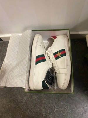 Gucci  sneakers - Säljer mina Gucci sneakers i bra skick. Skorna har snörning, rund tå och platt sula. Röd och grön detalj på hälen. Levereras med originalkartong och påse.