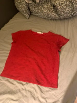 Röd glittrig topp från KappAhl - Snygg röd topp från KappAhl med glittrigt tyg och korta ärmar. Enkel och rak modell som passar perfekt till jeans eller kjol. T-shirten har rund halsringning och är riktigt bekväm att bära.