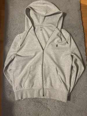 Ljusgrå hoodie från Polo Ralph Lauren - Hej! Säljer nu min ljusgrå zip hoodie från Polo Ralph Lauren med dragkedja framtill, två fickor och klassisk huva. Liten broderad logga på bröstet. Perfekt för en chill och stilren look. Denna kofta är väldigt eftertraktad och jag känner att den använts nog av mig därav säljer jag den. Inga klara tecken på användning och materialet är lika bra som när jag köpte den! Priset är ej hugget i sten och vid frågor så är det bara att ställa!