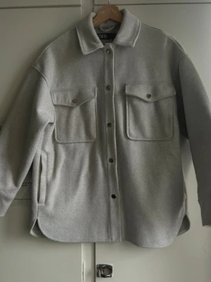 Skjortjacka - Zara - Säljer en beige/ljusgrå overshirt från Zara. Modellen är varm, mjuk och perfekt som både jacka och skjorta. Den har fyra fickor: två stora bröstfickor med lock och knappar samt två sidfickor som är diskreta och praktiska.  Overshirten har en lätt oversized passform, rak modell och tryckknappar framtill. Passar både till vardag och lager-på-lager. Rosa - grå färg  Skick: Mycket fint skick, sparsamt använd. Storlek: XS