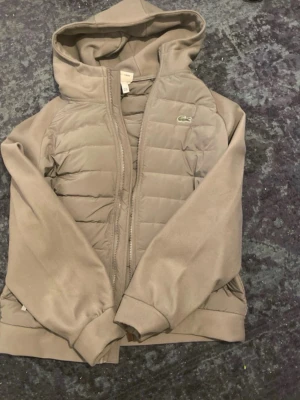 Mörk grå vindjacka från Lacoste - Snygg mörk grå vindjacka från Lacoste med huva och dragkedja framtill. Jackan har quiltade partier på bröstet och det klassiska krokodilmärket på vänster sida. Tillverkad i polyester och har långa ärmar samt ribbade muddar. Oanvänd och redo för skick🌀