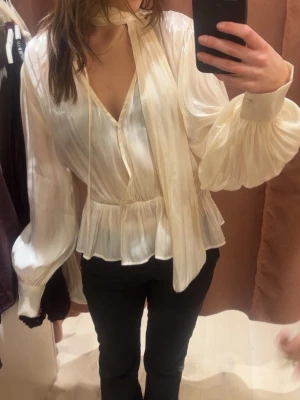 Krämvit blus med knytband - Snygg krämvit blus i glansigt material med volang nedtill och knytband vid halsen. Aldrig använd med prislappen kvar💕