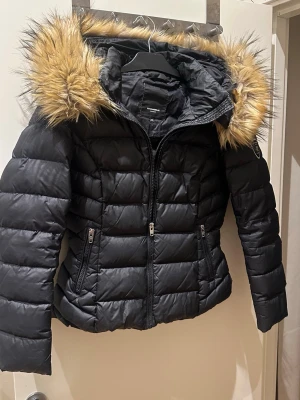 Svart dunjacka med fuskpäls - Snygg svart dunjacka med fluffig fuskpäls runt huvan och quiltad design. Jackan har dragkedja framtill, två fickor med dragkedja och en figurnära passform. Perfekt för kalla dagar och ger en trendig look.