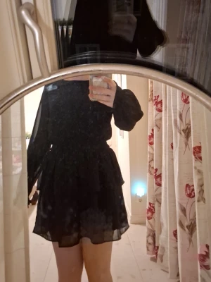 Svart kort klänning från Kappahl - Säljer en svart kort klänning från Kappahl med långa puffärmar och volangdetaljer i kjolen. Klänningen har en flowy passform och är tillverkad i ett tunt, lätt material som ger en snygg siluett.