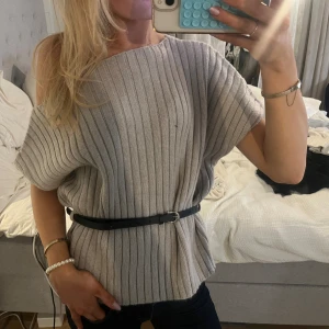 Ribbad grå stickad topp med bälte - Säljer en ribbad stickad topp i grå med korta ärmar och rund hals. Toppen har ett svart bälte i midjan som ger en snygg siluett. Perfekt att styla med jeans eller kjol för en trendig look.