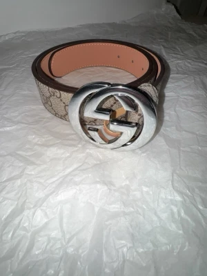 bälte beige - Snyggt bälte i beige och brunt med klassiskt mönster och stor silverfärgad dubbel spänne. Bältet är gjort i canvas och läder, perfekt för att lyfta vilken outfit som helst.