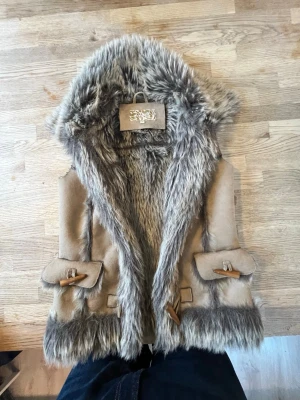 Beige pälsväst, stl 34 - Snygg beige pälsväst med mjuk fuskpäls i gråbrunt och dekorativa träknappar. Västen har stora fickor framtill och bred krage i pälsimitation. Perfekt för lager-på-lager och ger en cool boho-vibe.