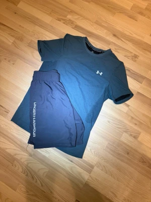 Under Armour träningsset M - Säljer ett träningsset från Under Armour med en mörkgrön t-shirt och svarta shorts. T-shirten har rund hals och logga på bröstet, shortsen har resår i midjan och vit logga längs benet. Perfekt för gym eller löpning.