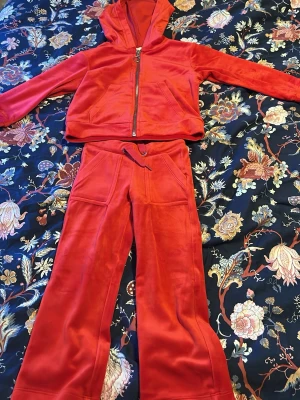 Rött velourset med hoodie och byxor - Säljer ett mjukt och skönt set i röd velour med zip-hoodie och matchande byxor. Hoodien har dragkedja och fickor framtill, byxorna har dragsko i midjan och vida ben samt fickor både fram och bak. Perfekt för en avslappnad och trendig look.