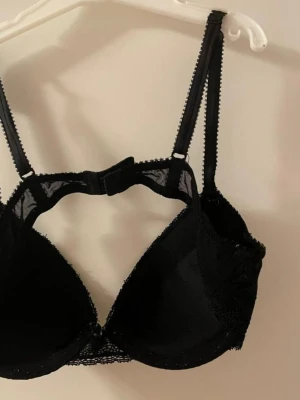 Svart push-up bh med spetsdetaljer storlek 70A - Snygg svart push-up bh med tunna axelband och fina spetsdetaljer längs kanterna. Bh:n har bygel och vadderade kupor för extra stöd och form. Klassisk knäppning bak och justerbara band för bästa passform. Saknar etiketter.
