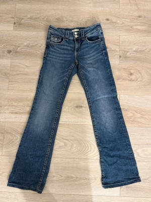 Blå bootcut jeans med dubbla knappar - Snygga blå bootcut jeans med dubbla knappar i midjan och klassiska fem fickor. Jeansen har en lätt tvättad look och raka ben som går ut något nedtill. Perfekta för en avslappnad och trendig stil.