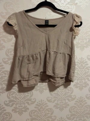 Beige blus/bluzka - Söt beige blus från Gina Tricot med volang nedtill och v-ringning. Ärmarna är korta och dekorerade med spetsdetaljer som ger en romantisk känsla. Perfekt att styla med jeans eller kjol för en avslappnad look.