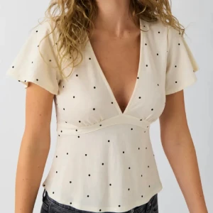 Prickig v-ringad blus med knyt - Säljer en ljus, prickig blus med v-ringning och korta, lite fladdriga ärmar. Toppen har ett fint knytband i ryggen som ger en snygg siluett. Materialet känns mjukt och lätt, perfekt för varmare dagar. Prickarna är svarta och ger en cool kontrast mot den ljusa bakgrunden. Superfin och populär!