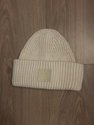  Acne Studios mössa - Säljer en beige stickad mössa från Acne Studios med det klassiska ansiktsloggan framtill. Mössan är ribbstickad och tillverkad i 100% ull, perfekt för att hålla värmen under kyliga dagar. Enkel och stilren design som passar till allt.