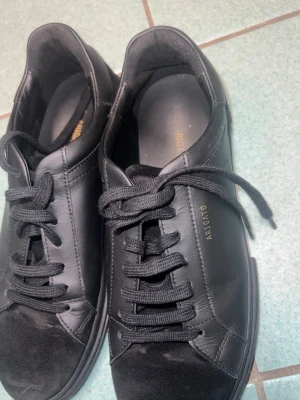 Svarta sneakers från Axel Arigato - Snygga svarta sneakers från Axel Arigato med stilren design. Skorna har rund tå, platt sula och snörning. Ovandelen är i skinn med detaljer i mocka på tån och hälen. Diskret logga i guld på sidan. Perfekta för dig som gillar minimalistisk stil.