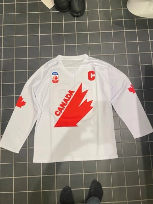 Vit hockeytröja Canada Gretzky 99 - Säljer en vit hockeytröja med röd lönnbladsgrafik och texten CANADA på framsidan. Tröjan har långa ärmar med röda lönnblad på båda sidor, röd kaptensmärke 'C' och Labatt-logga. På ryggen står det GRETZKY och siffran 99 i rött.