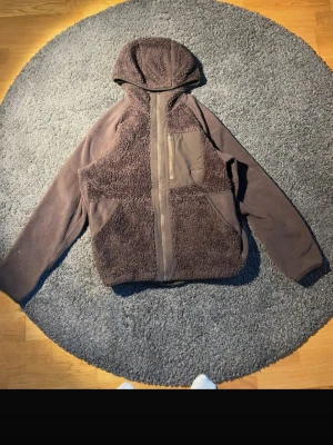 Mörkbrun fleecejacka med huva från Uniqlo - Mysig mörkbrun teddyjacka med huva och hel dragkedja framtill. Jackan har en stor bröstficka med dragkedja och två sidofickor. Materialet är fluffigt teddytyg kombinerat med slät polyester på fickan och dragkedjan. Perfekt för kyliga dagar och chill stil.
