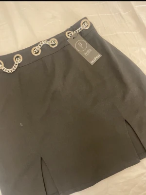 Svart kortkjol med kedjedetalj Boohoo - Snygg svart kortkjol från Boohoo Petite med silverfärgade kedjor och metallöljetter i midjan. Kjolen har två slitsar framtill och dragkedja i sidan för enkel påklädning. 